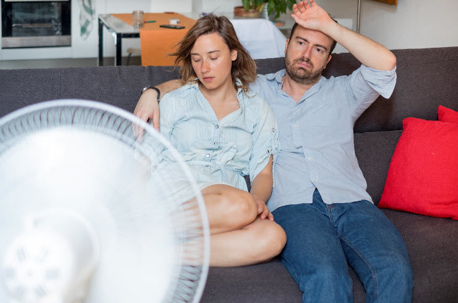 A distressed man and woman sit on a couch, visibly hot, while a fan attempts to cool them.