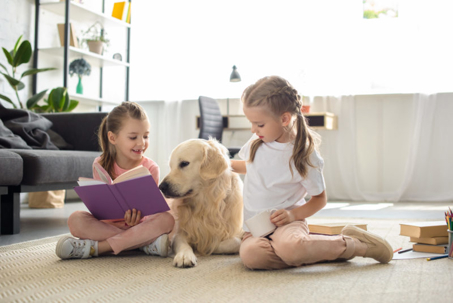 Two young girls and a golden retriever dog relax on a living room rug.