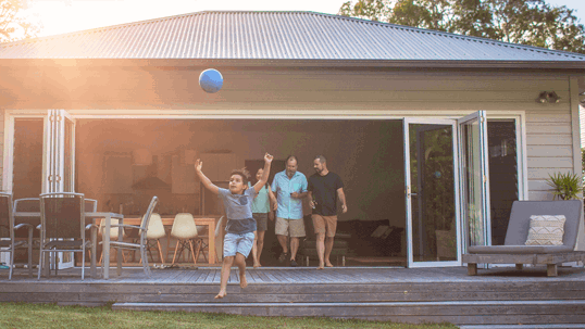 A boy jumps for a ball on a sunny lawn as a family exits a modern house with open doors onto a deck, enjoying their summer day.