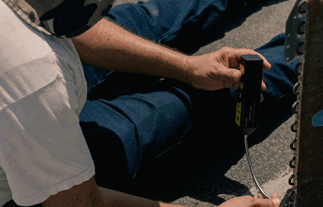 A person in blue jeans and a white shirt works on dark ground outdoors, holding a black tool with a partial yellow logo near a metal object.