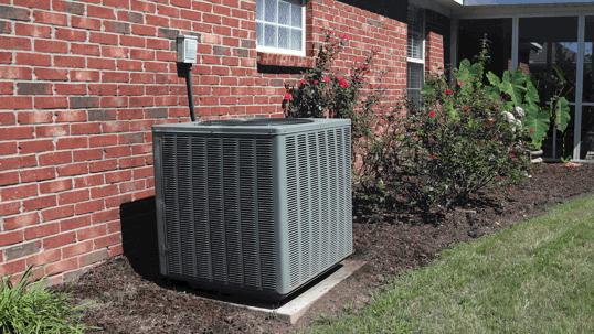 An outdoor air conditioning unit sits next to a brick house. It is surrounded by fresh mulch and green landscaping under a bright sky.