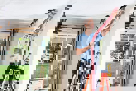 A technician on a ladder cleans an air duct in a modern home's ceiling with a red hose.