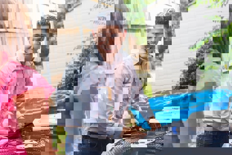 A smiling HVAC technician, wearing sunglasses and holding a clipboard, talks to a female homeowner next to an outdoor air conditioning unit.