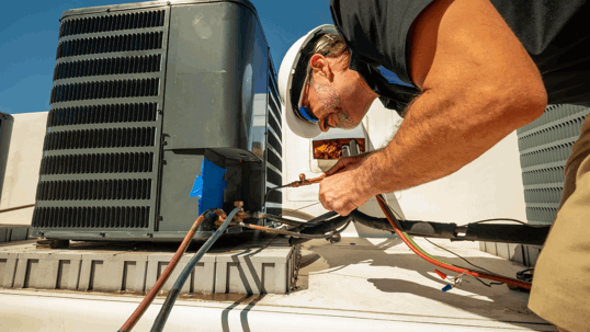 A technician in a hard hat and safety glasses works on an outdoor HVAC unit, connecting copper lines on a rooftop.