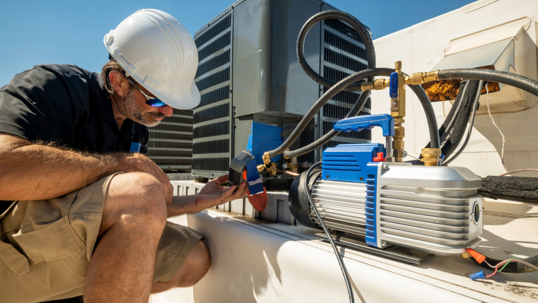 An HVAC technician in a hard hat services a rooftop unit with a vacuum pump.