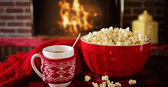 A cozy scene with a mug in a red knitted cozy and a red bowl of popcorn by a fireplace.