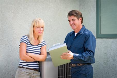 A male technician with a clipboard consults a smiling woman by an outdoor AC unit
