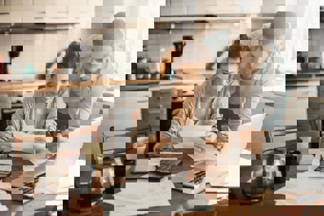 A couple sits at their kitchen table, reviewing documents and a laptop.