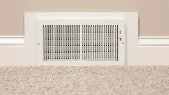 A white rectangular HVAC return air vent is installed in a beige wall above light-colored carpet.