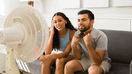 A man and woman sit on a couch, feeling hot in front of a white standing fan.