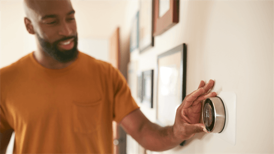 A smiling man in an orange shirt adjusts a smart thermostat on a wall.
