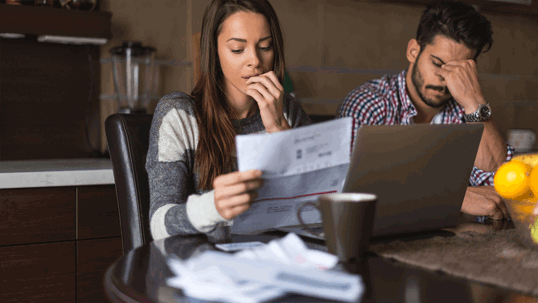 A stressed couple sits at a table, reviewing documents and a laptop.