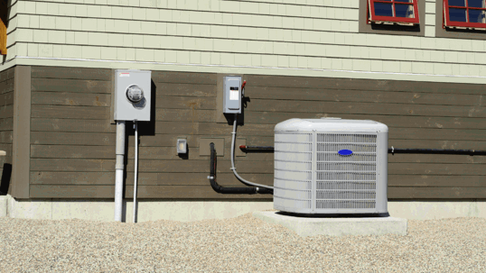 An outdoor AC unit and electrical boxes against a building with light and dark siding, set on gravel.