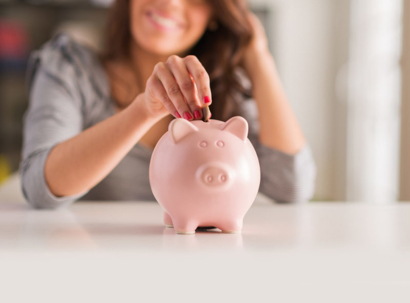 A smiling woman places a coin into a pink piggy bank on a white table