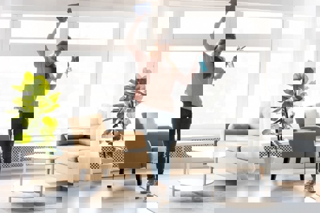 A young woman happily dances with a cleaning cloth and spray bottle in a sunlit living room