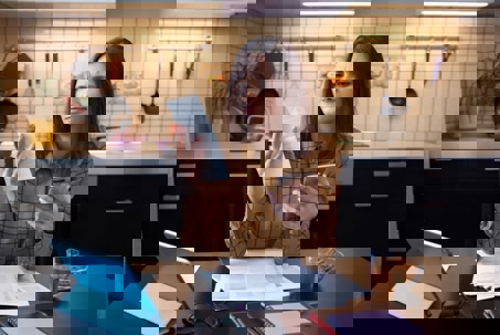 A woman with a perplexed expression holds a bill and smartphone at her kitchen table